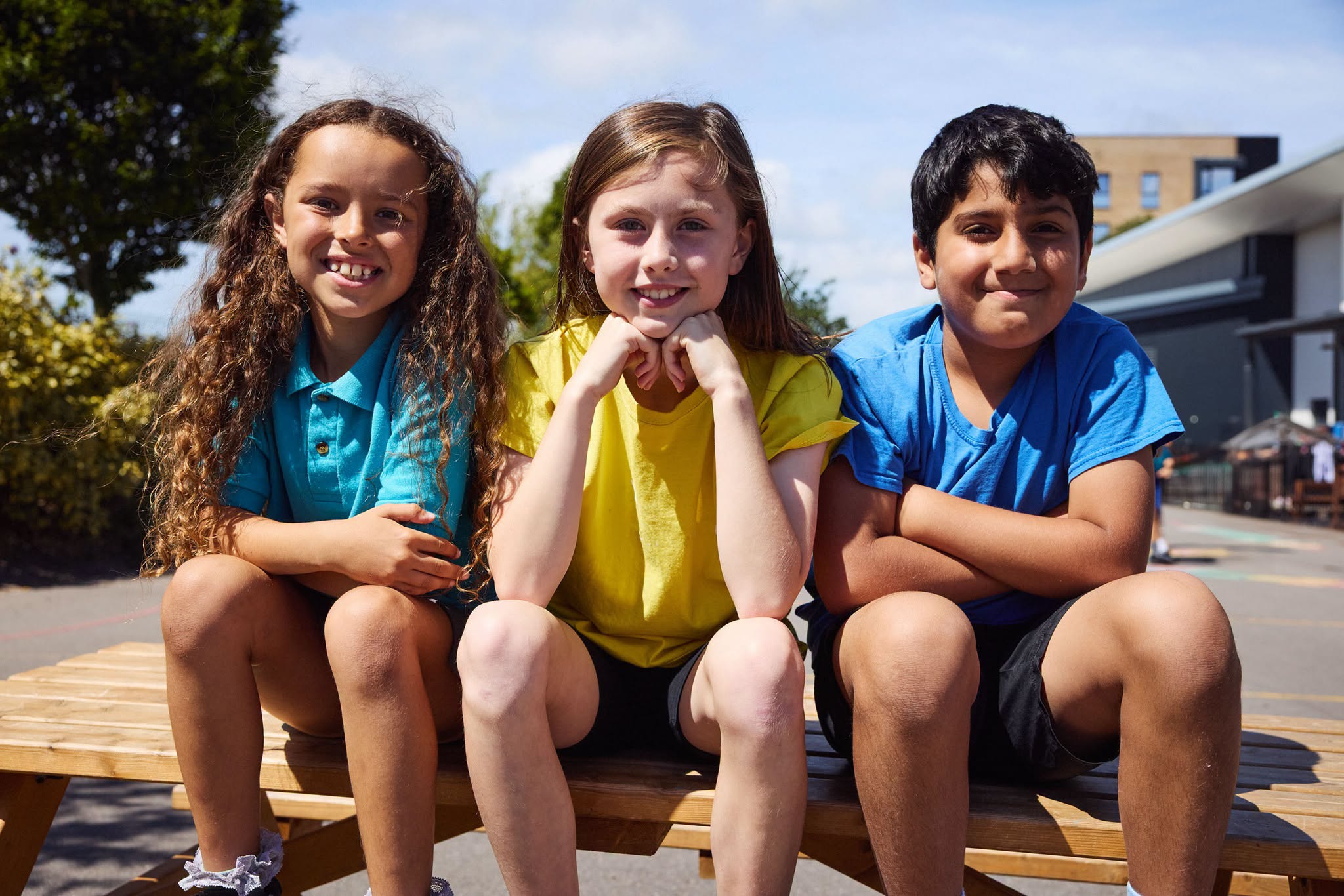A group of primary school-aged children sitting together outdoors on a wooden bench in a school playground or educational setting.