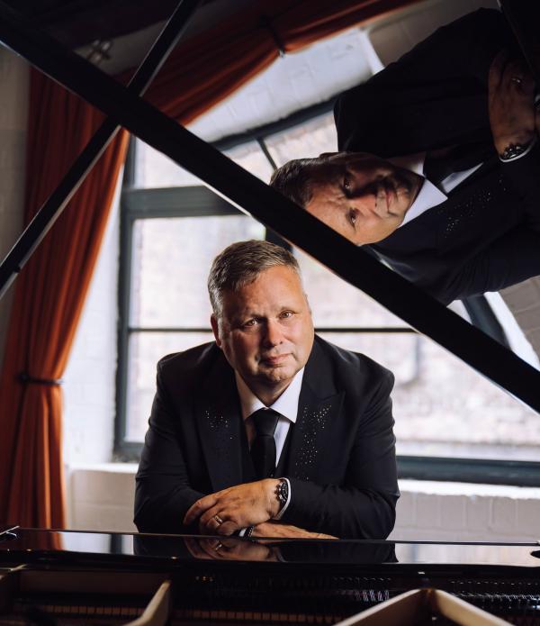 Portrait of singer Paul Potts leaning on a grand piano, with his reflection visible in the raised piano lid and large windows in the background.