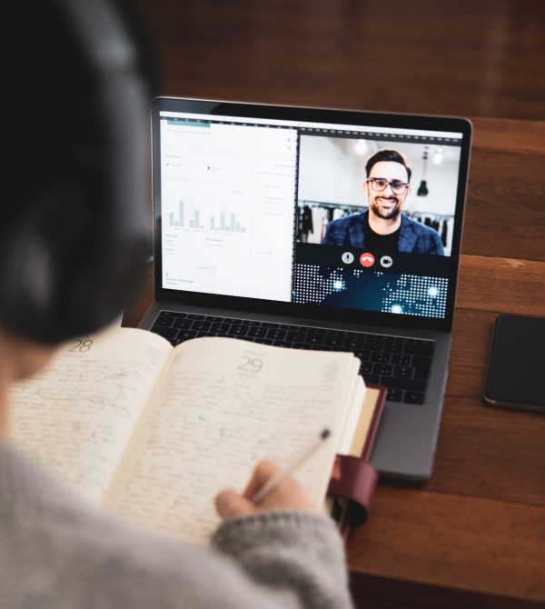 Person wearing headphones taking notes in a notebook while participating in a video call on a laptop; the screen shows charts and graphs alongside the video feed.