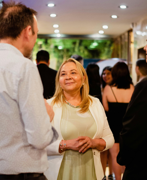 A woman in a pale green top and a cream cardigan talks to a tall man in a light blue shirt at a networking event, against a backdrop of people talking to each other. There is a green wall and spotlights in the ceiling.