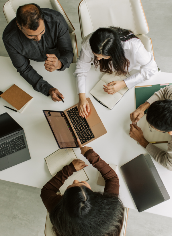 Group of people with laptops working at a white desk in an office. All are focused on one laptop, suggesting collaborative work.