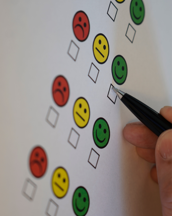 A hand marking a checkbox next to a green smiling face on a customer satisfaction survey or feedback form. The scale ranges from red (negative) to green (positive) emoji faces.