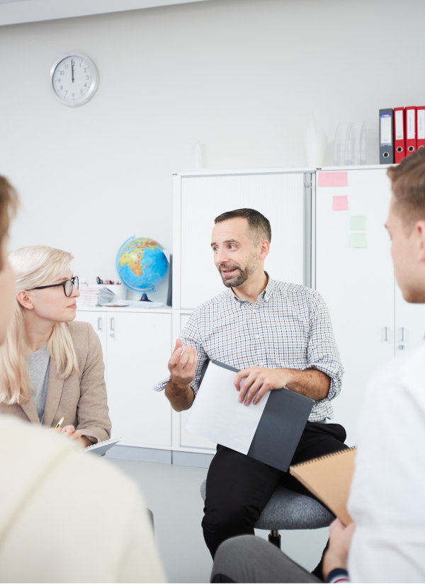 A collaborative team discussion where a speaker is presenting ideas and documents to his colleagues, seated together in a bright office space.