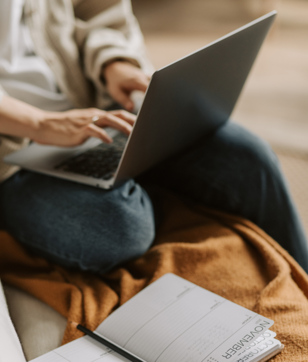 Person sitting on a couch with a laptop on their lap, typing. An open planner showing the month of November and a pen rest on a brown blanket beside them.