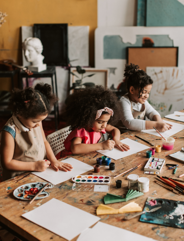 Children seated at a large wooden table engaged in an art activity, with blank sheets of paper, watercolor palettes, paintbrushes, and various art supplies spread across the table. The background shows an art studio with paintings, canvases, and sculpting materials.