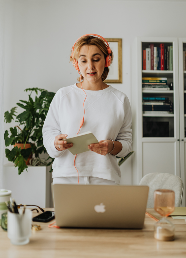 Person standing at a desk with an open laptop, holding a notebook and wearing headphones. The desk has various items including a glass, a candle, and a container with pens. In the background, there is a bookshelf and a large green plant.