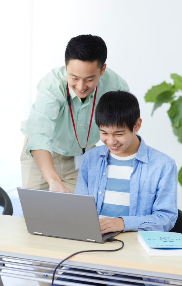 Two people in a bright indoor setting, one seated at a desk using a laptop while the other stands nearby pointing at the screen; a book is placed on the desk next to the laptop.