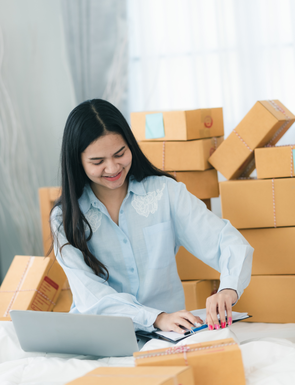 Person sitting indoors surrounded by multiple cardboard boxes, working on a laptop and writing in a notebook.