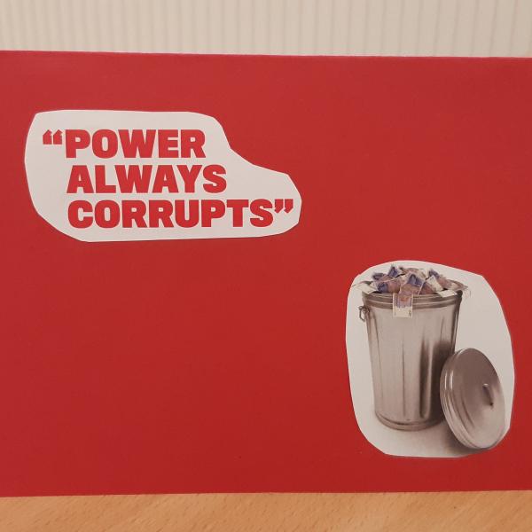 A red card with the words 'power always corrupts' and a bin full of money