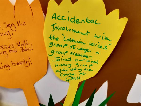 A yellow cut-out flower with the handwritten words ‘Accidental involvement with the ‘Lothian Voices’ group. Through a group member joined Oor Mad History group after doing the course at QMU’