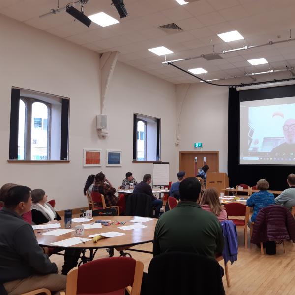 A group of people in a large room watching a presentation on a screen.