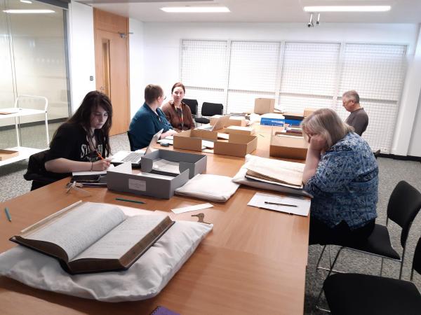 A group of people sitting around a table, studying materials, there is an open book resting on a pillow in the front of the picture.