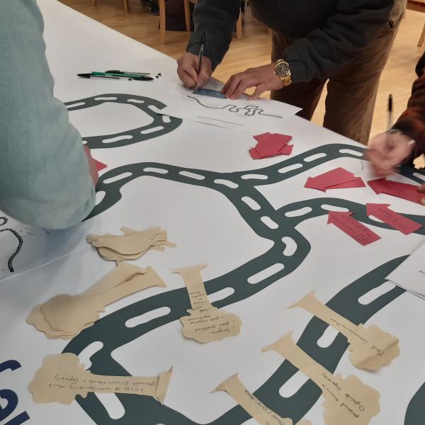 A sheet of paper with a graphic of a road, peoples hands are seen writing on smaller pieces of paper.