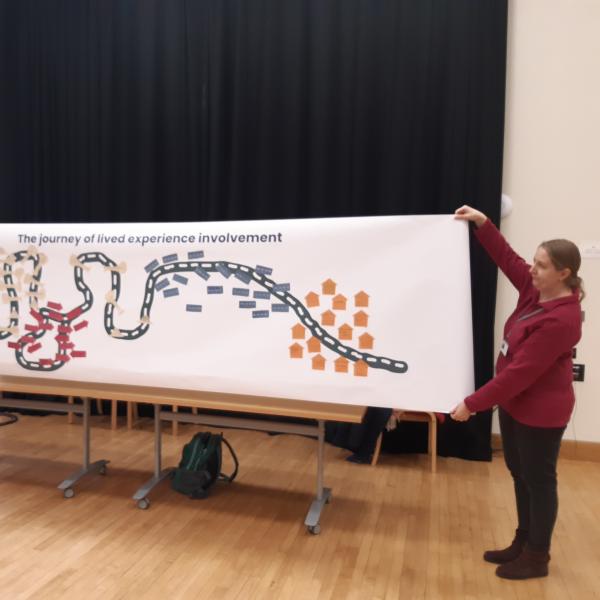 A person holding a large scroll with graphics of a road and people's thoughts about the journey of lived experience.