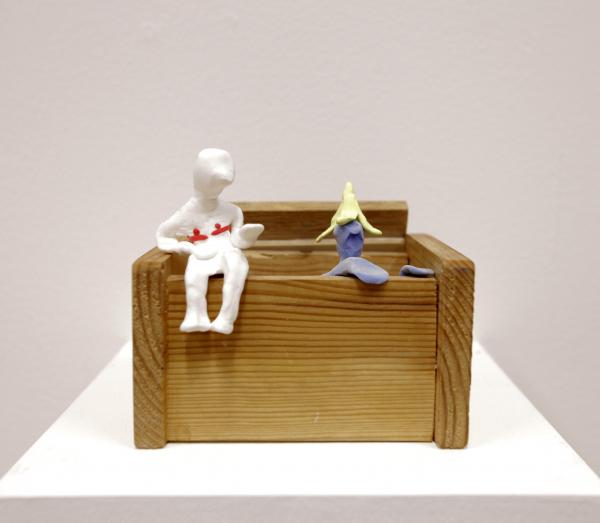 An artwork that shows a wooden box with two sculpted figures climbing out of it, one is white, the other purple