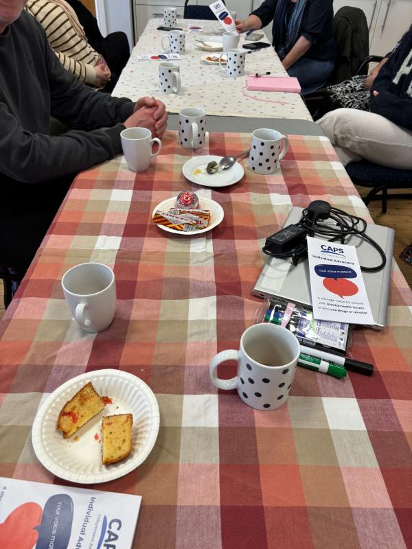 A brightly coloured table top with mugs, plates and CAPS leaflets, people’s legs and arms can be seen as they sit around the table. 