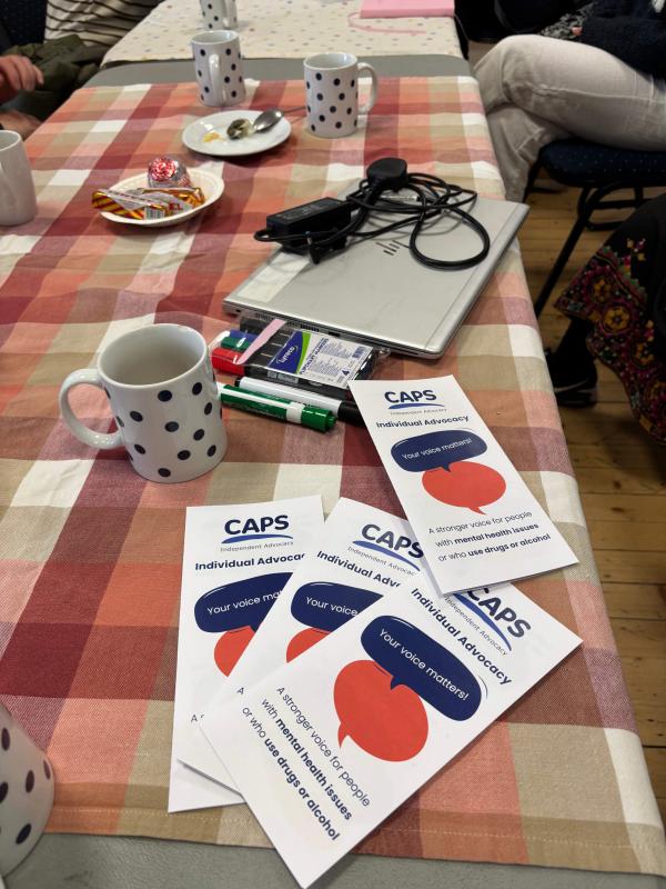 A brightly coloured table top with mugs, plates and CAPS leaflets, people’s legs and arms can be seen as they sit around the table. 