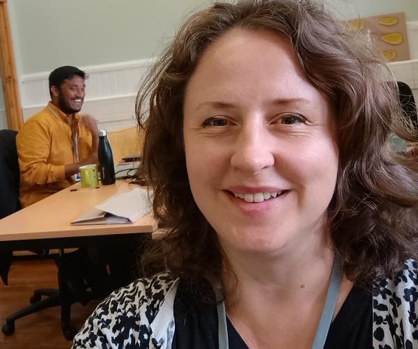 A picture of Catherine smiling. An office with other workers is in the background.