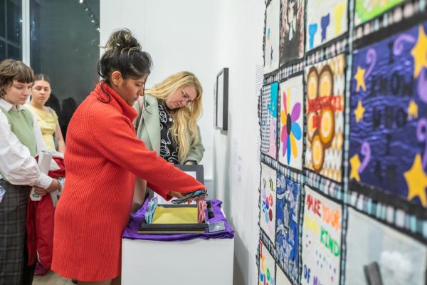 A range of artworks in bright colours on a wall with a group of people stand looking at an artwork in the form of a book