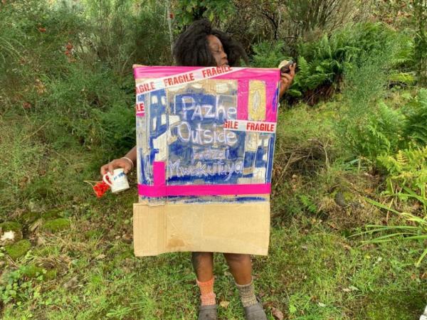 A woman, Fadzai Mwakutuya, stands in a grassy landscape wearing an artwork made from card that reads Pazhe Outside Fadzai Mwakutuya