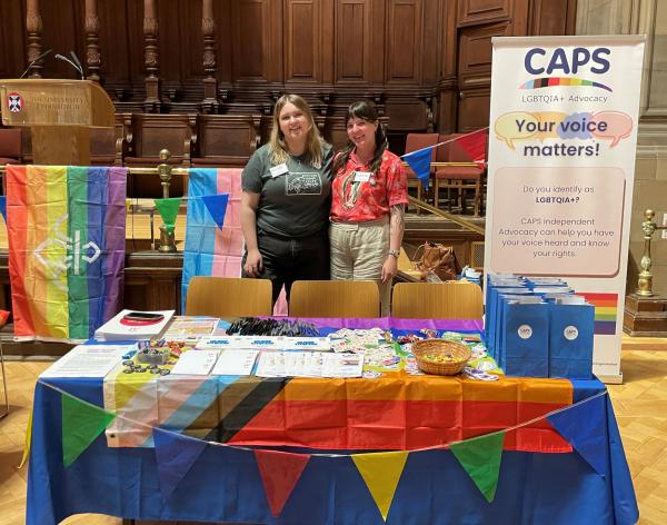 Two people standing behind a stall set out with leaflets and pride flags, a banner in the background reads 'Your Voice Matters' 