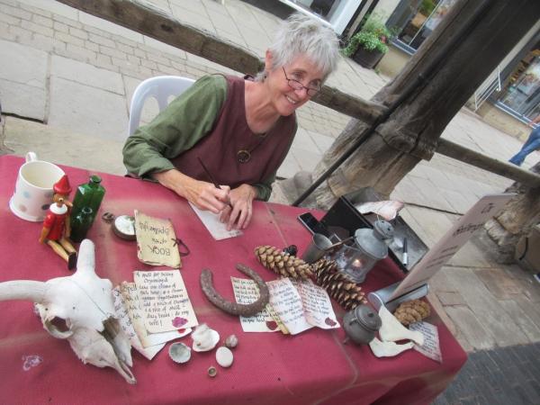 Spellwright Jean Atkin, sat at a desk, surrounded by lucky symbols, writing a spell on parchment