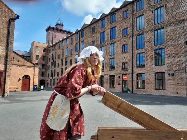 A young woman dressed in costume, in front of a brick mill building