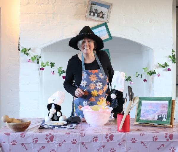 A woman wearing black clothes and an apron, she is holding a whisk. On a table in front of her is a mixing bowl and two cat puppets