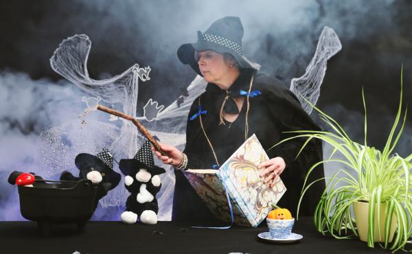 A woman dressed as a witch, holding a wand. Next to her are two cat puppets, both wearing witch hats