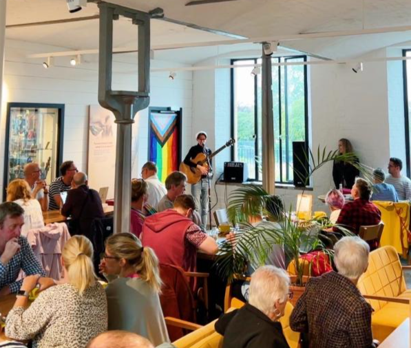 A busy cafe, with a man stood in the corner holding an acoustic guitar. The pride progress flag is on display behind him