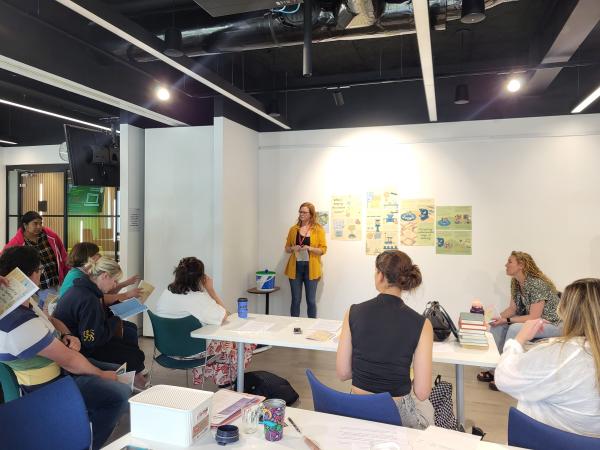 Woman at the front of the room presenting to people sitting at desks.