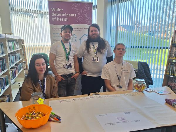 Co-researchers gathered around a desk at an event. The background is in a library setting with books on shelves, there is also a 'wider determinants of health' roller banner behind the group.