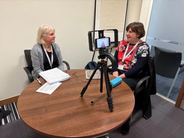 Two women sat at a table having a conversation in front of a video recording device set up on a tripod.