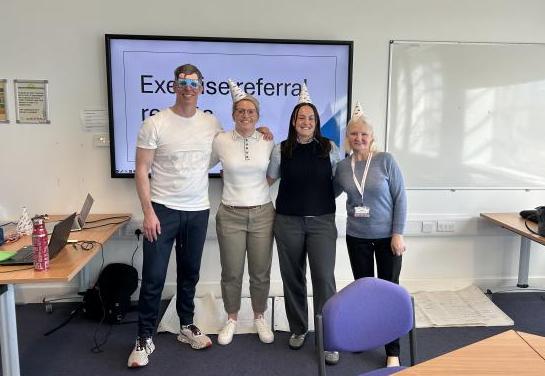 One man and three women posing for a photo in front of a presentation. They are wearing party hats.