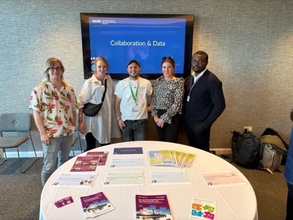 Group photo of colleagues at an event stood behind a table. The screen behind the shows a slide with 'collaboration and data' on the screen.