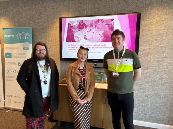 Two men and a woman stood in front of a powerpoint screen at an event.