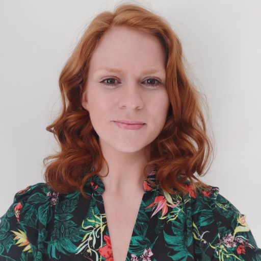Woman with red hair facing the camera wearing a green shirt with a bright coloured pattern.