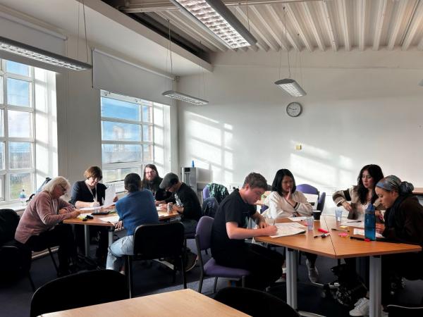 Image of attendees on a training course. Participants are of mixed ages and ethnicity, sat around two tables working.