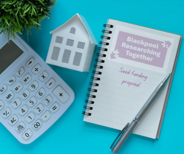 Notepad showing seed funding proposal, next to a pen, calculator, white house and plant. 