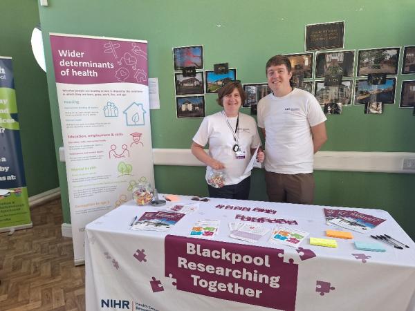 Blackpool Researching Together team at an event. Two people stood behind a table and pop up banner containing informative leaflets and images to update on the work of Blackpool Researching Together.