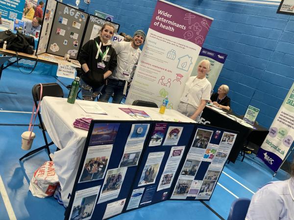 Blackpool Researching Together team at an event. Two people stood behind a table and pop up banner containing informative leaflets and images to update on the work of Blackpool Researching Together.