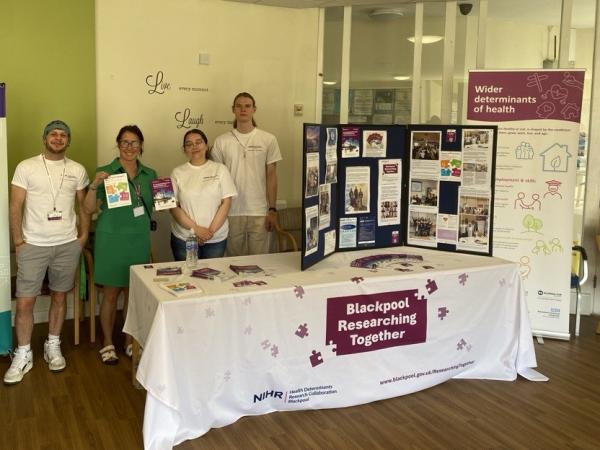 Blackpool Researching Together team at an event. Four people gathered around a table containing informative leaflets and images to update on the work of Blackpool Researching Together.