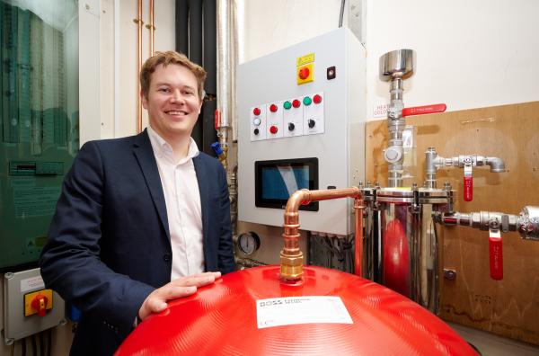 Councillor Jack Hemingway standing in front of a control panel for the heating system, resting his hand on a red storage tank