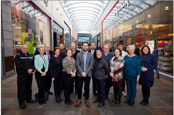 A photo of 15 people, including Councillor Michael Graham and other members of the City Centre Management Board, stood in the middle of Trinity Walk shopping centre