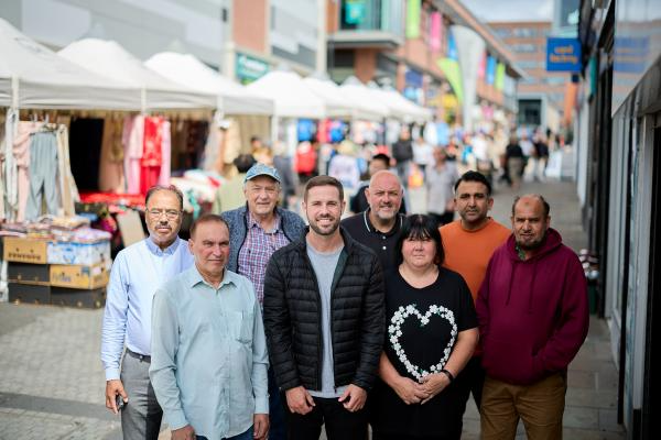 A photo of Councillor Michael Graham smiling with seven happy market traders in front of market stalls