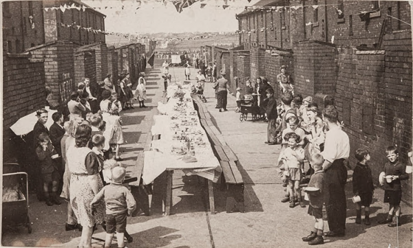 A black and white photograph of a street party gathering celebrating VE Day.