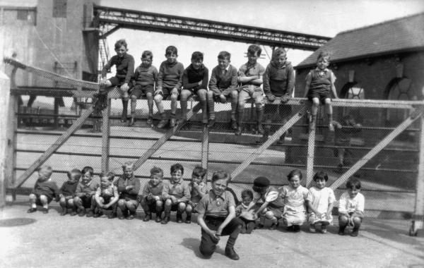 An old photograph of 8 children sat on a gate and 15 children sat in front of the gate outside a colliery