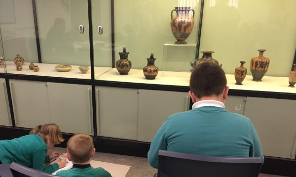 children drawing Greek pots next to cabinet of Greek pots on display