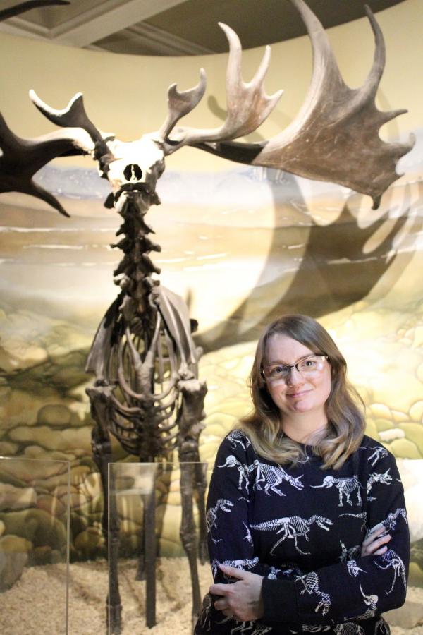 young museum staff member in dinosaur jumper stands in front of a Elk skeleton