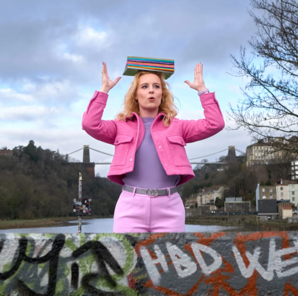 a woman in a pink jacket stands with Clifton suspension bridge in background. She has books on her head